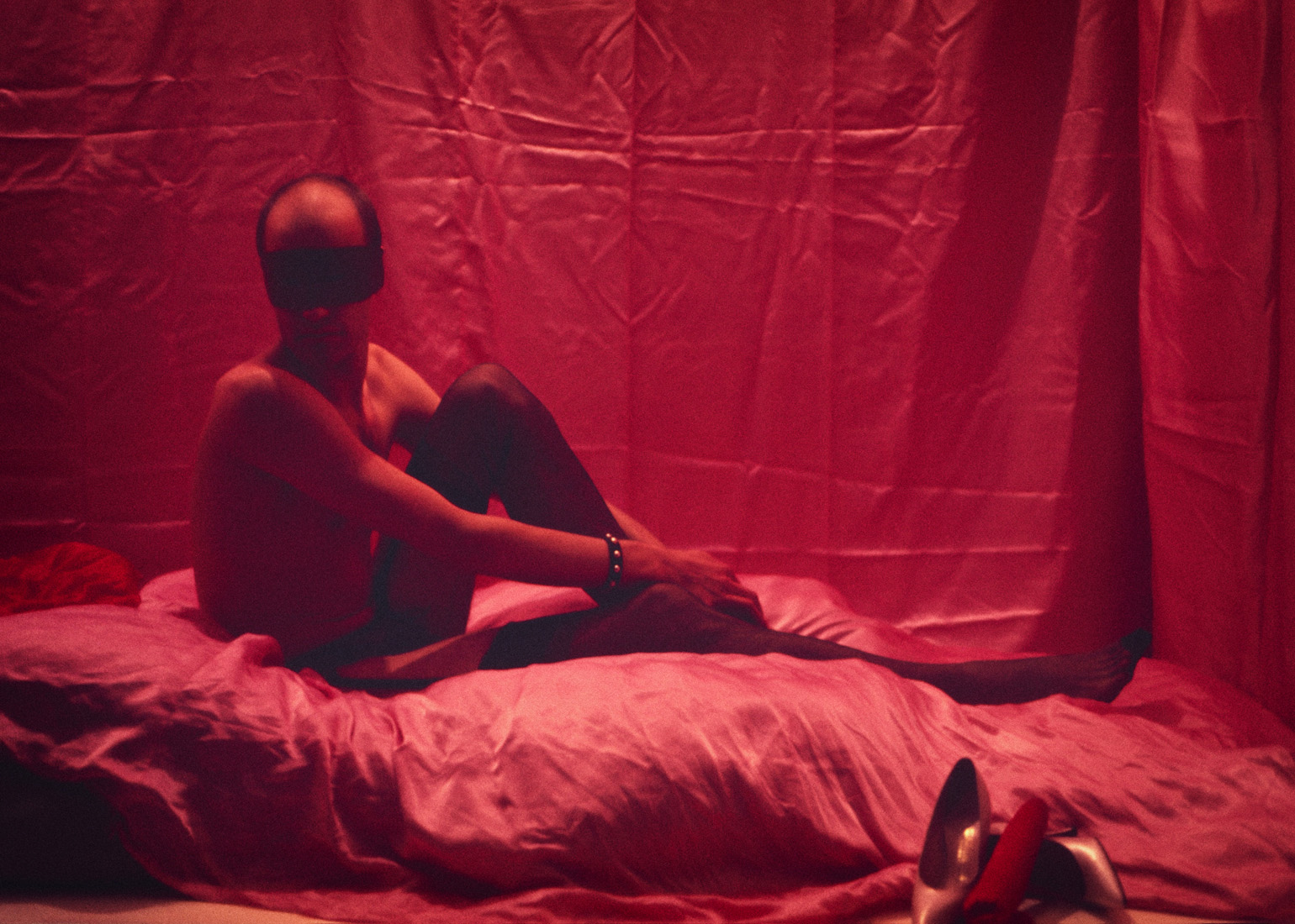 A masked figure posing on pink satin sheets